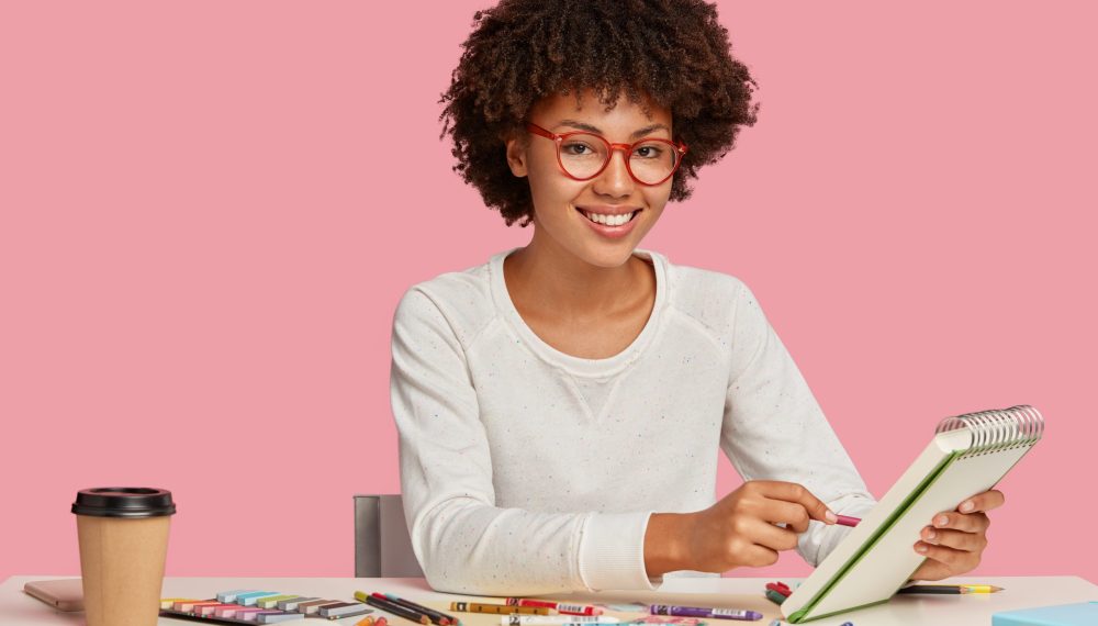 Horizontal shot of cheerful Afro African painter wears red rim spectacles, makes sketches in spiral notebook with pencil, enjoys aromatic beverage, surrounded with stationary, smiles positively Horizontal shot of cheerful Afro African painter wears red rim spectacles, makes sketches in spiral notebook with pencil, enjoys aromatic beverage, surrounded with stationary, smiles positively