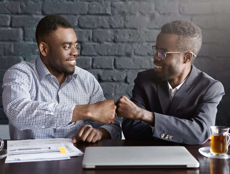 Business, teamwork and collaborataion concept. Two dark-skinned businesspeople in formal wear giving each other fist bump after successful agreement at meeting, happy with signing profitable contract